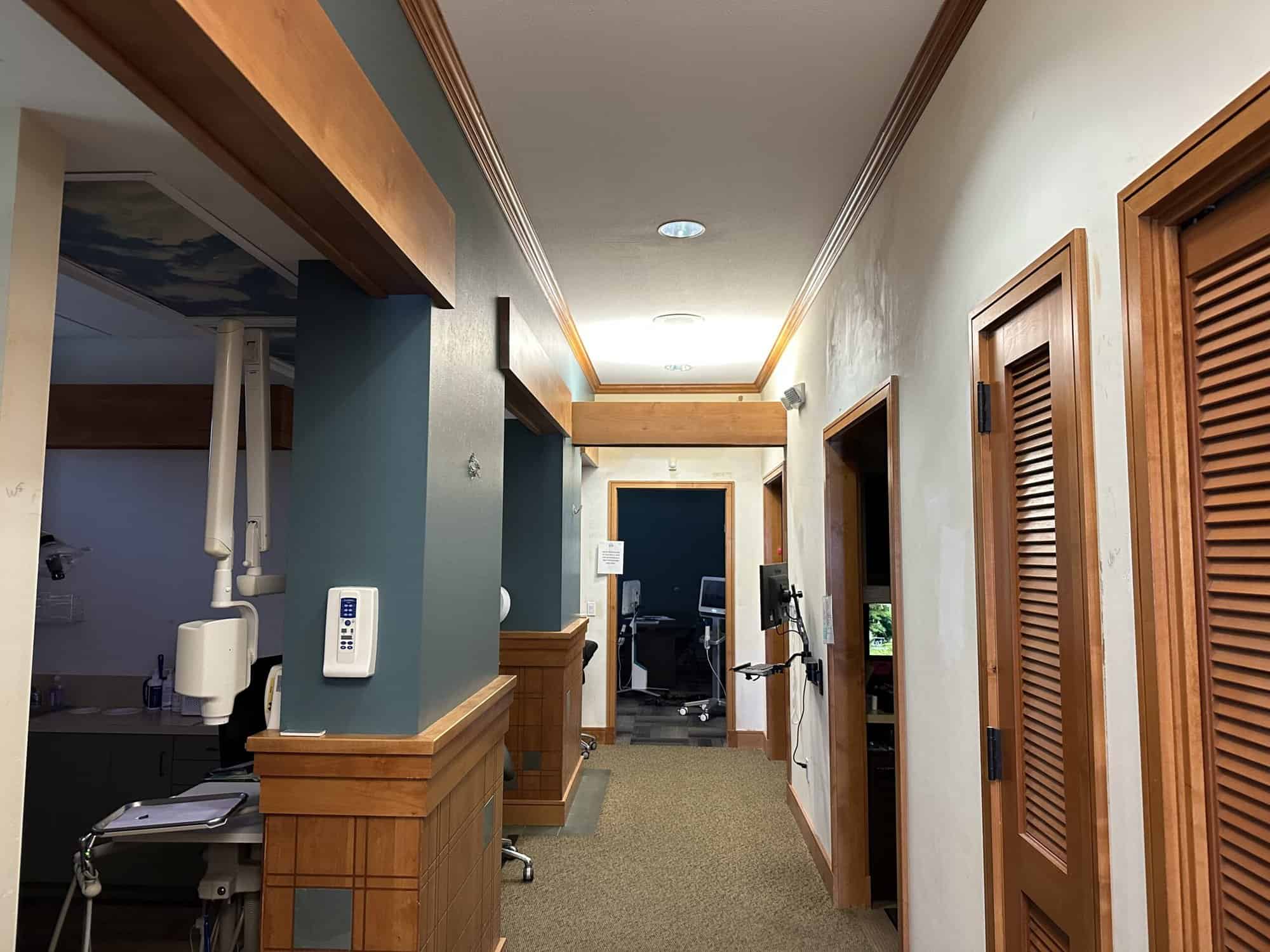 A long, narrow hallway in a medical facility, with several doors leading to different rooms. The walls are painted in a light blue color, and there is a wooden trim along the ceiling and baseboards.