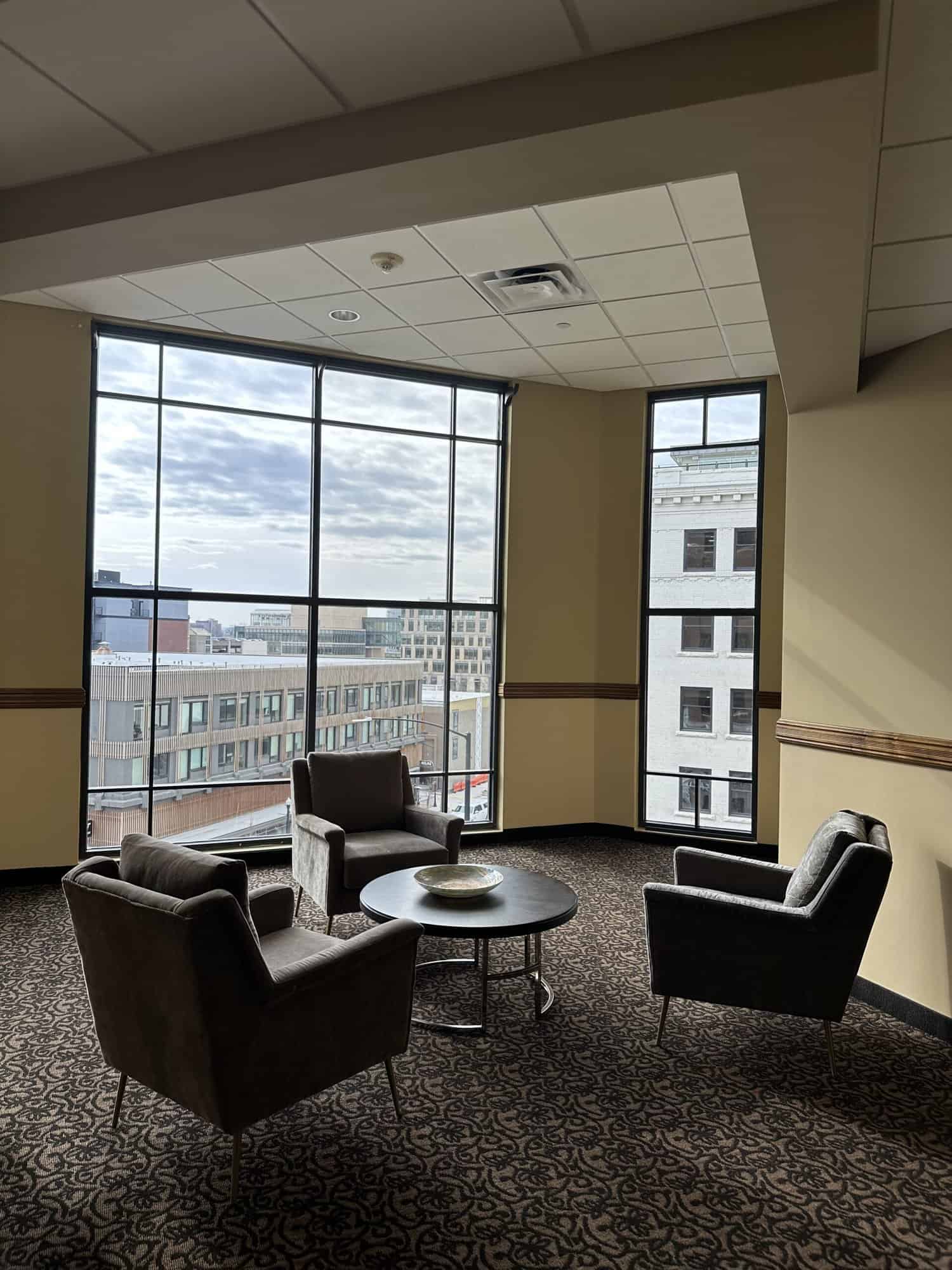 A spacious lounge area with large windows offering a view of the city skyline. Comfortable armchairs are arranged around a coffee table, creating a cozy and inviting atmosphere.