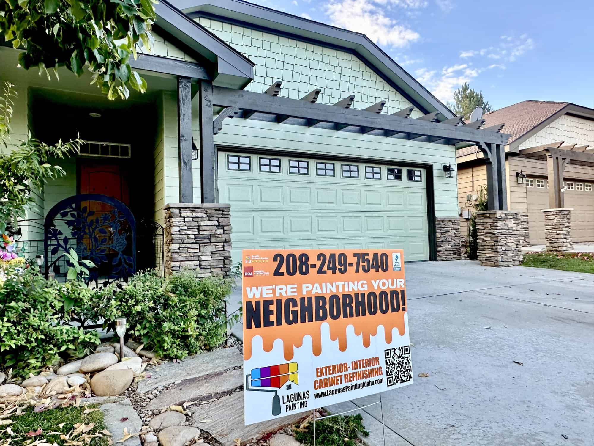 A modern, one-story house with a green exterior and a stone accent wall. The house features a two-car garage with a wooden pergola above, a front porch, and a well-manicured lawn. A sign in the front yard advertises painting services.