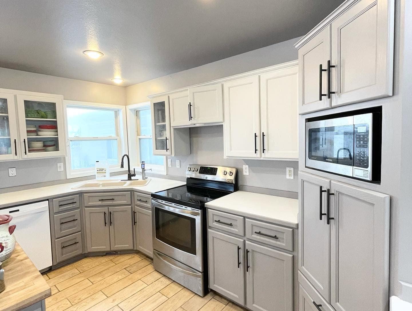 A modern kitchen with a mix of white and gray cabinets, black hardware, and stainless steel appliances. The kitchen features a large window, a tiled backsplash, and a wood-look floor.