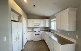 A kitchen with white cabinets, a white countertop, and a white refrigerator. The kitchen features a tiled floor, a window, and a light gray wall color.